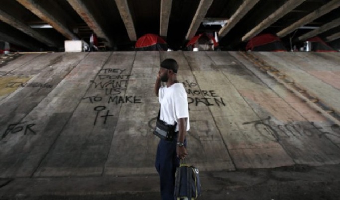 Un homme afro-américain sur la trentaine, pauvrement vêtu, un portable à l'oreille et traînant un trolley marche sous un pont en béton. Des tentes ont été dressées près des piliers. L'endroit est squalide, sale et plein de graffitis. 