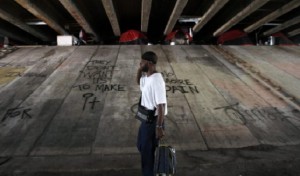 An afro-american man, a mobile phone to his ear and pulling a trolley, walks under the bridge at the Julia Tuttle Causeway. In the space between the bottom of the bridge and the top of its cement base, tents have been put in place. The ground is dirty and the cement covered in graffitis.